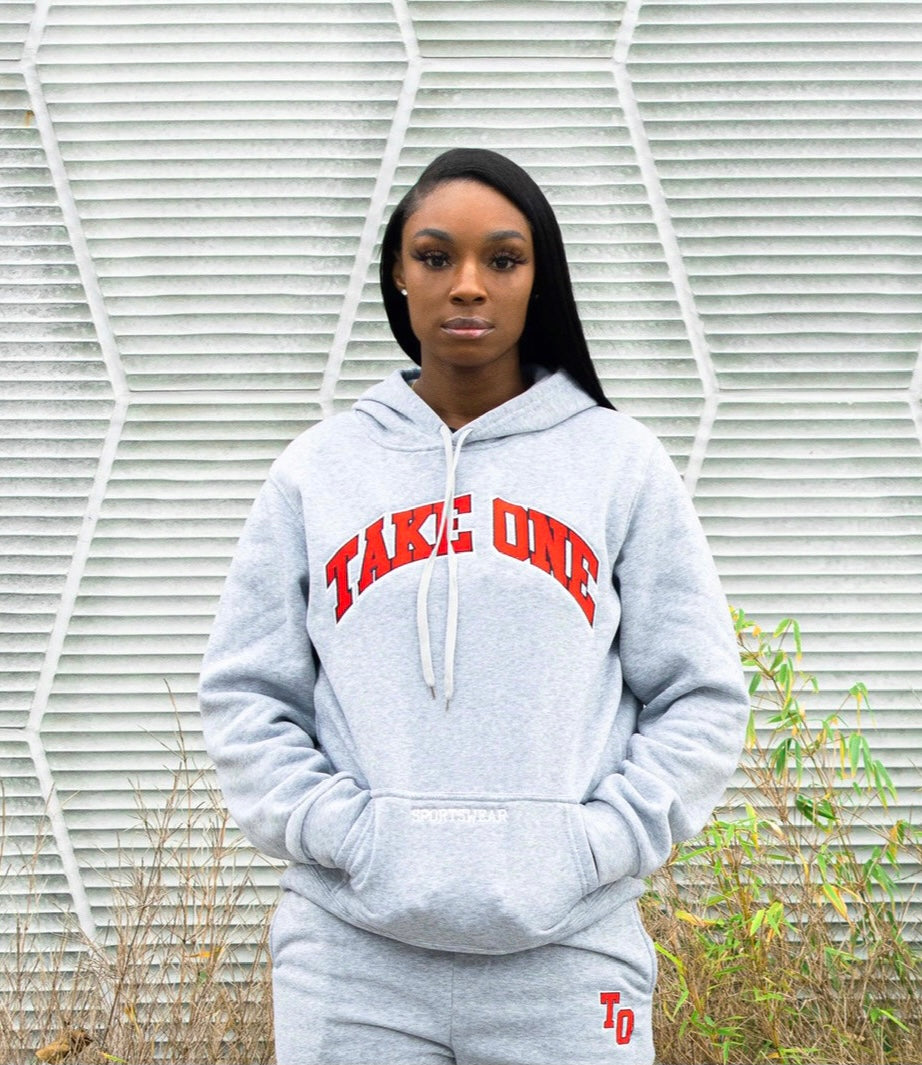 Woman modeling a heather grey Take One Sportswear Dynasty Hoodie with arched red TAKE ONE lettering, standing outdoors in matching grey sweatpants.