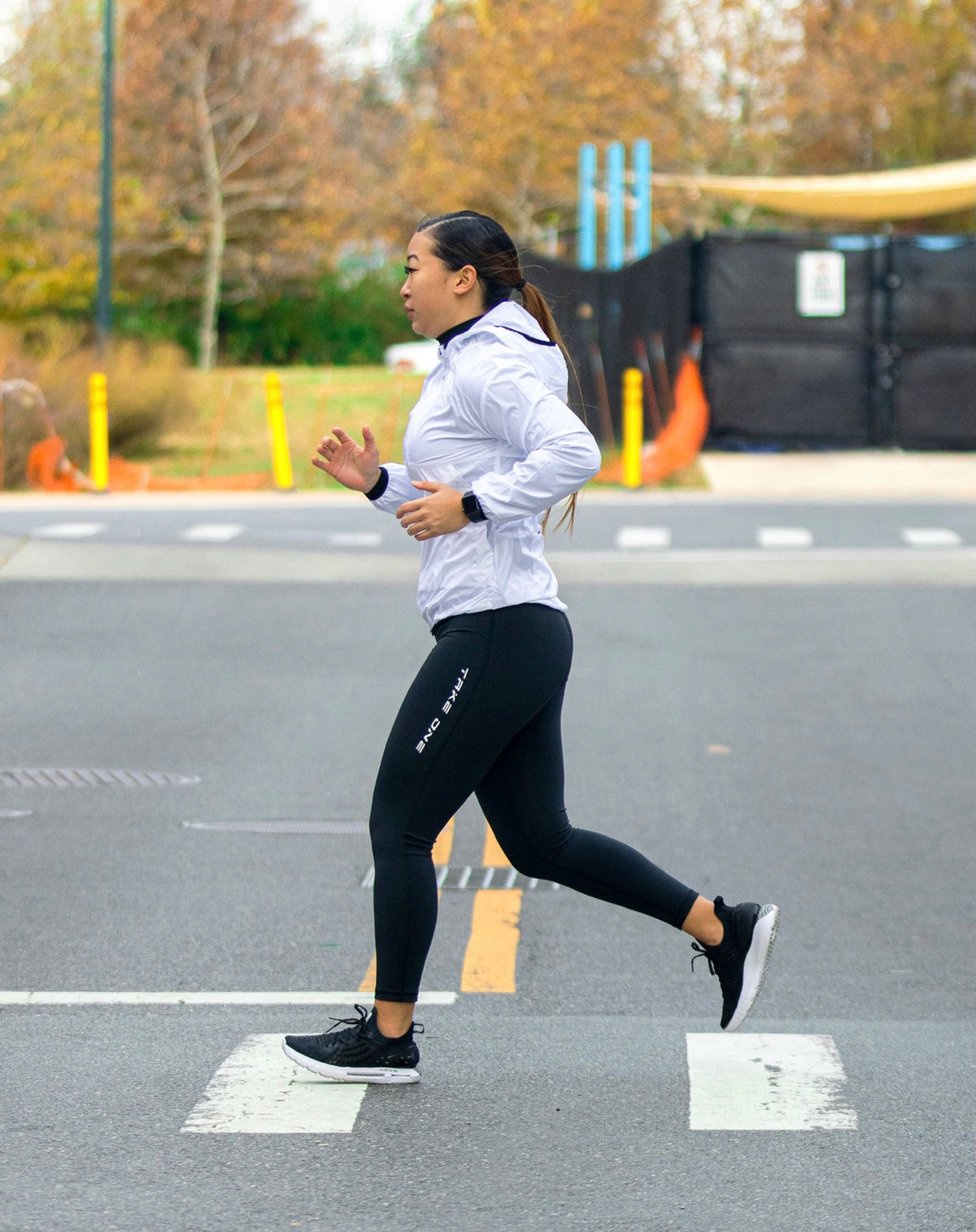 Woman running outdoors in black Take One Athena performance leggings, a light-colored athletic jacket, and black running shoes.