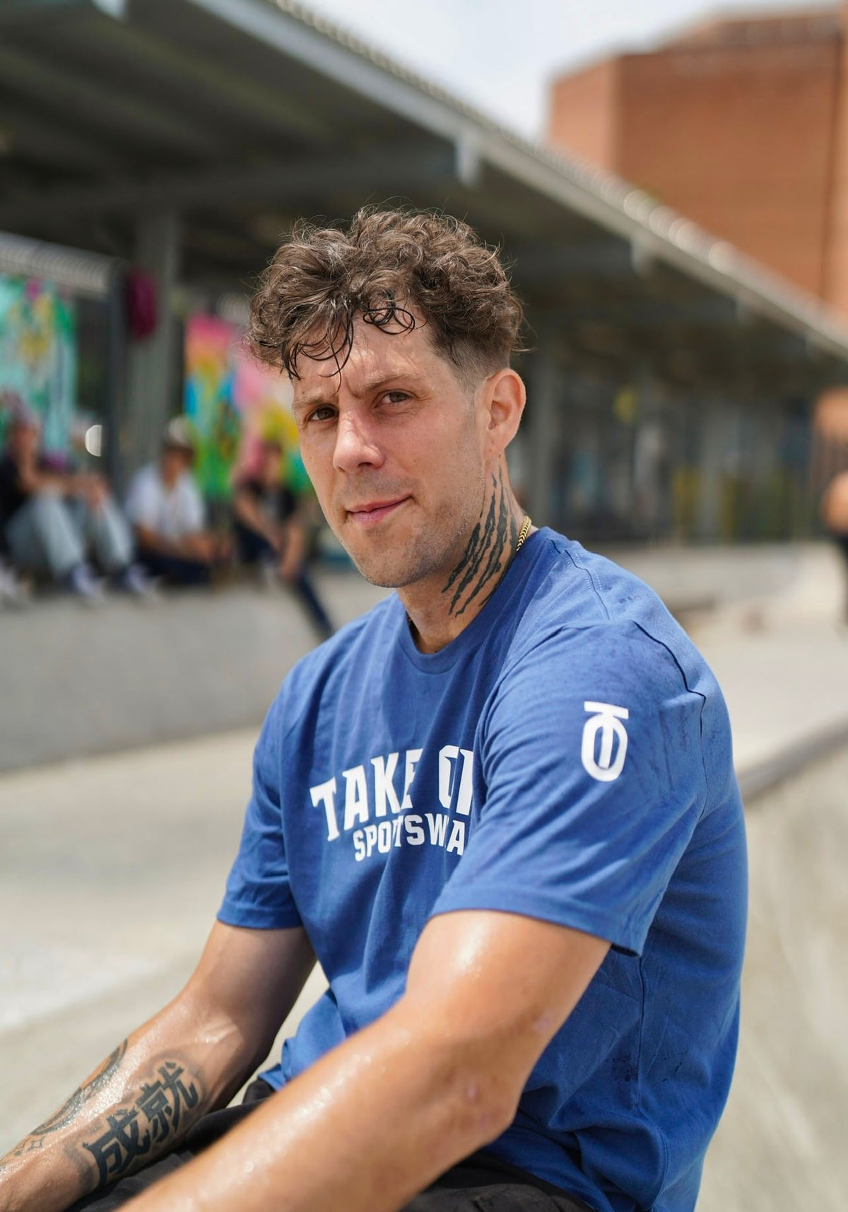 Male model seated outdoors in a blue Take One Chances Graphic T-Shirt with white chest lettering, styled with black shorts in an urban setting.