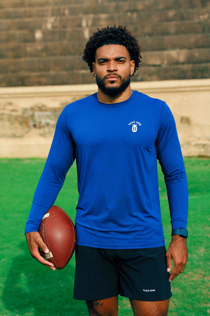 Male athlete wearing black Take One Sportswear Fleet Shorts with a blue long-sleeve training shirt while holding a football on a field.