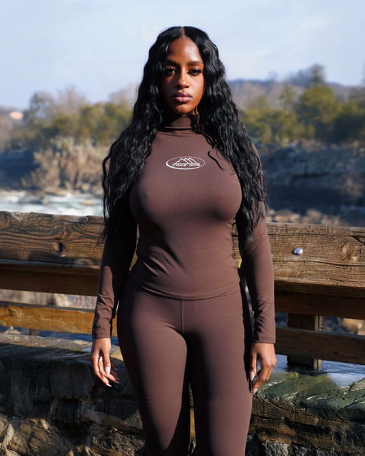 Model wearing a brown Pathfinder Axis fitted mock-neck long-sleeve top with matching leggings by Take One Sportswear, standing outdoors.