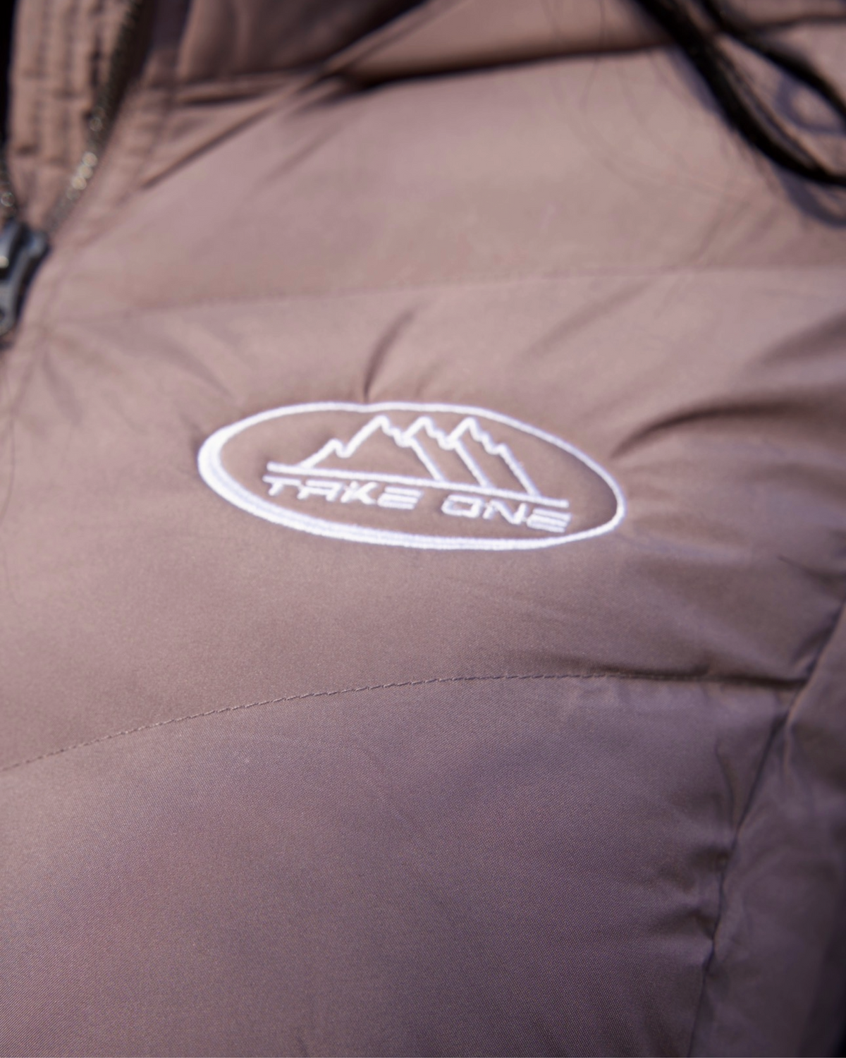 Close-up of the taupe Pathfinder Puffer Jacket by Take One Sportswear, showing the Trailway chest logo and quilted outer fabric.