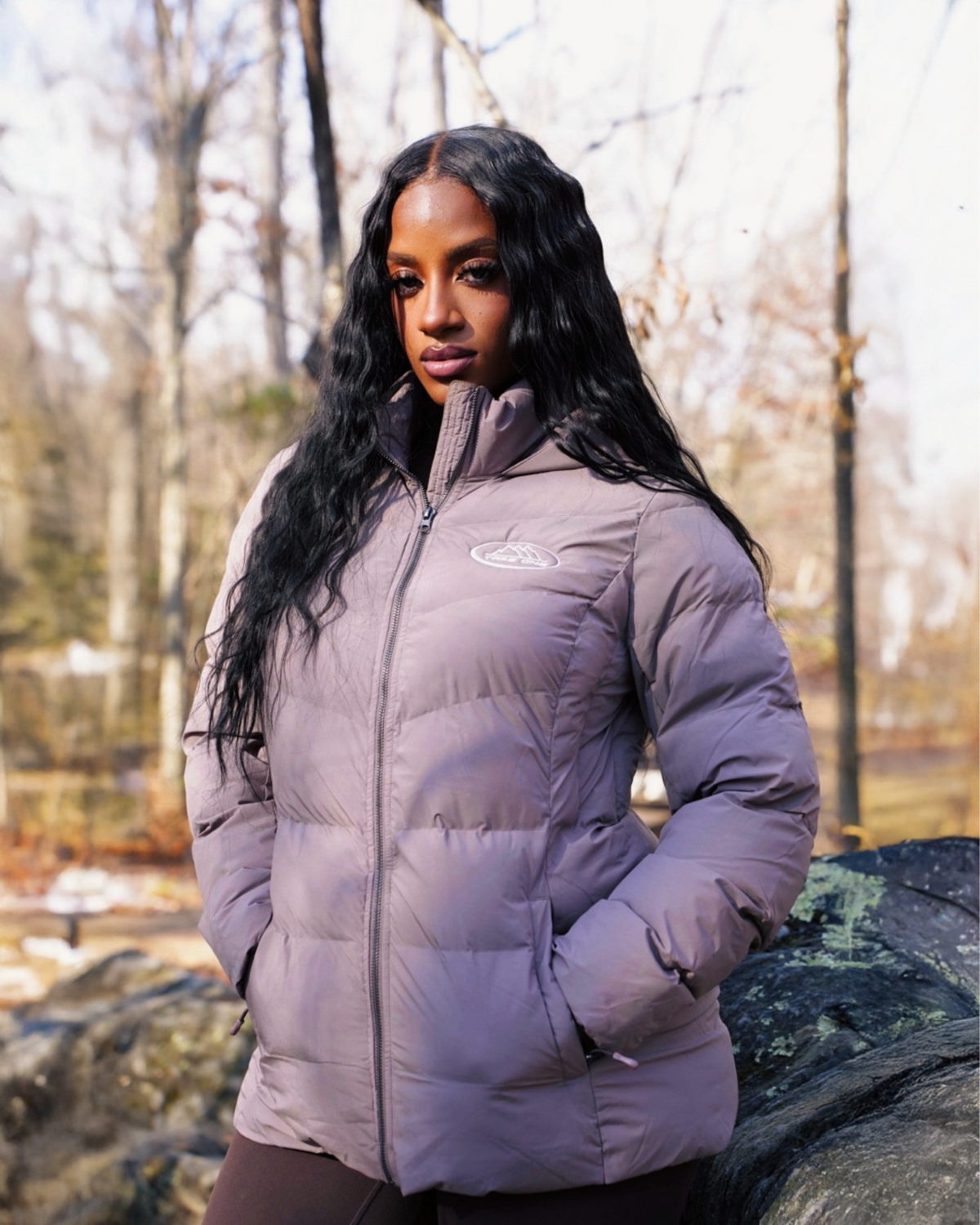 Model wearing a taupe Pathfinder Puffer Jacket by Take One Sportswear, with a high collar and quilted design, standing outdoors with hands in pockets.