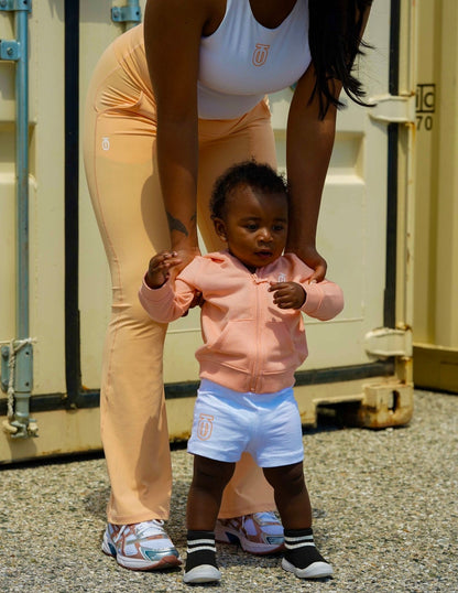 Toddler wearing light blue Take One Sportswear athletic shorts and a peach hoodie while being supported by an adult outdoors.