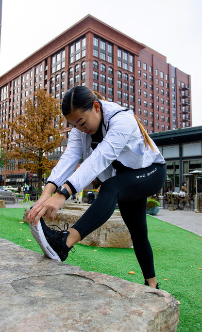 Woman wearing black Take One Sportswear Athena leggings while stretching outdoors beside a brick building.