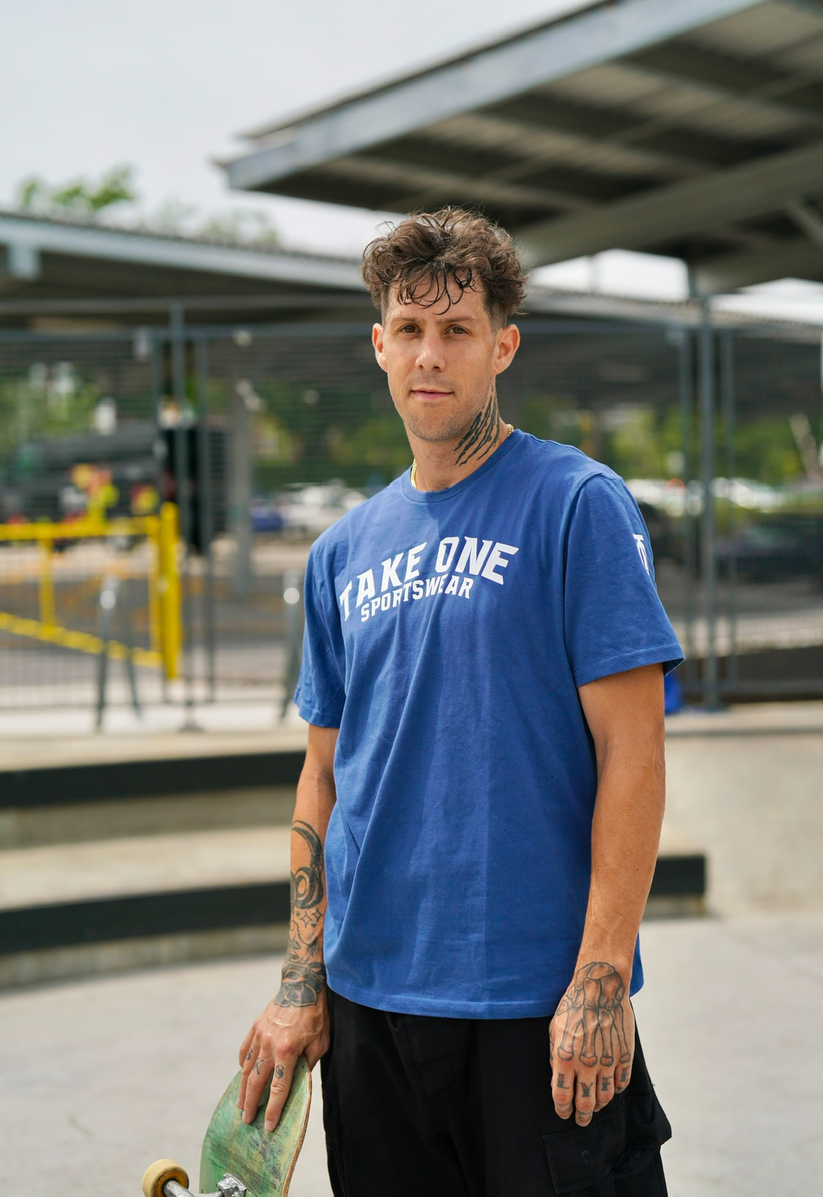Male model wearing a blue Take One Chances Graphic T-Shirt with white Take One lettering and black shorts while standing outdoors near an urban transit platform.