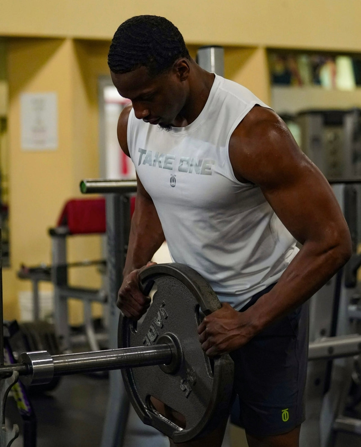 Male athlete wearing a white Take One Sportswear Dimension 2.0 Compression Tank while adjusting weight plates on a barbell inside a gym.
