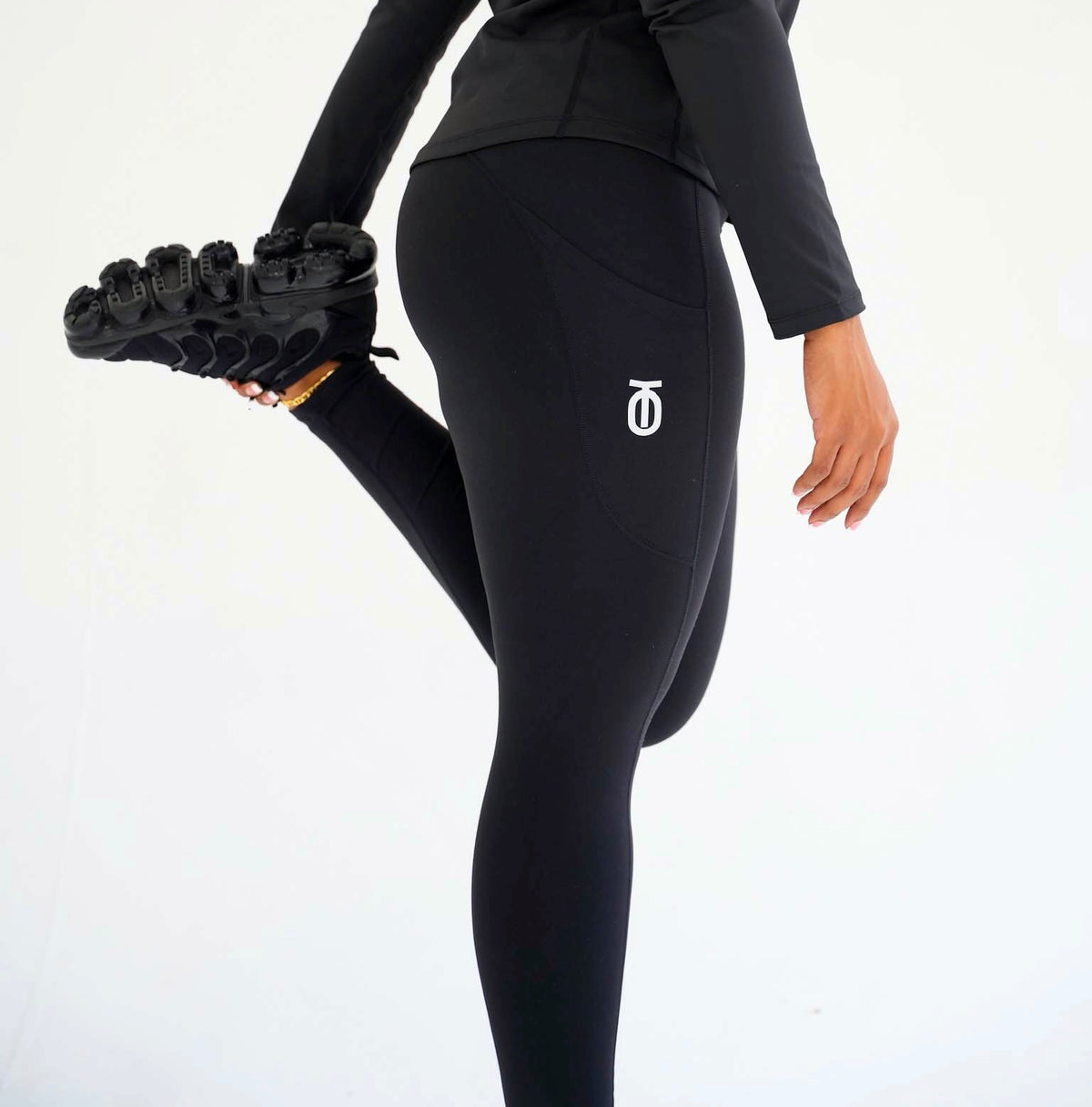 Side view of a woman stretching in black Take One Luxe Leggings, showing the fitted silhouette and small Take One logo near the hip.