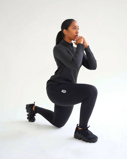 Woman performing a lunge in black Take One Sportswear Luxe performance leggings and a matching long-sleeve athletic top against a white studio background.