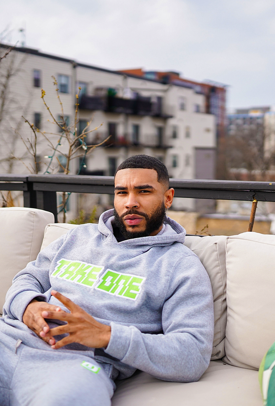 Man seated outdoors wearing a grey Take One Sportswear Prime Pullover Hoodie with a bright green Take One graphic.