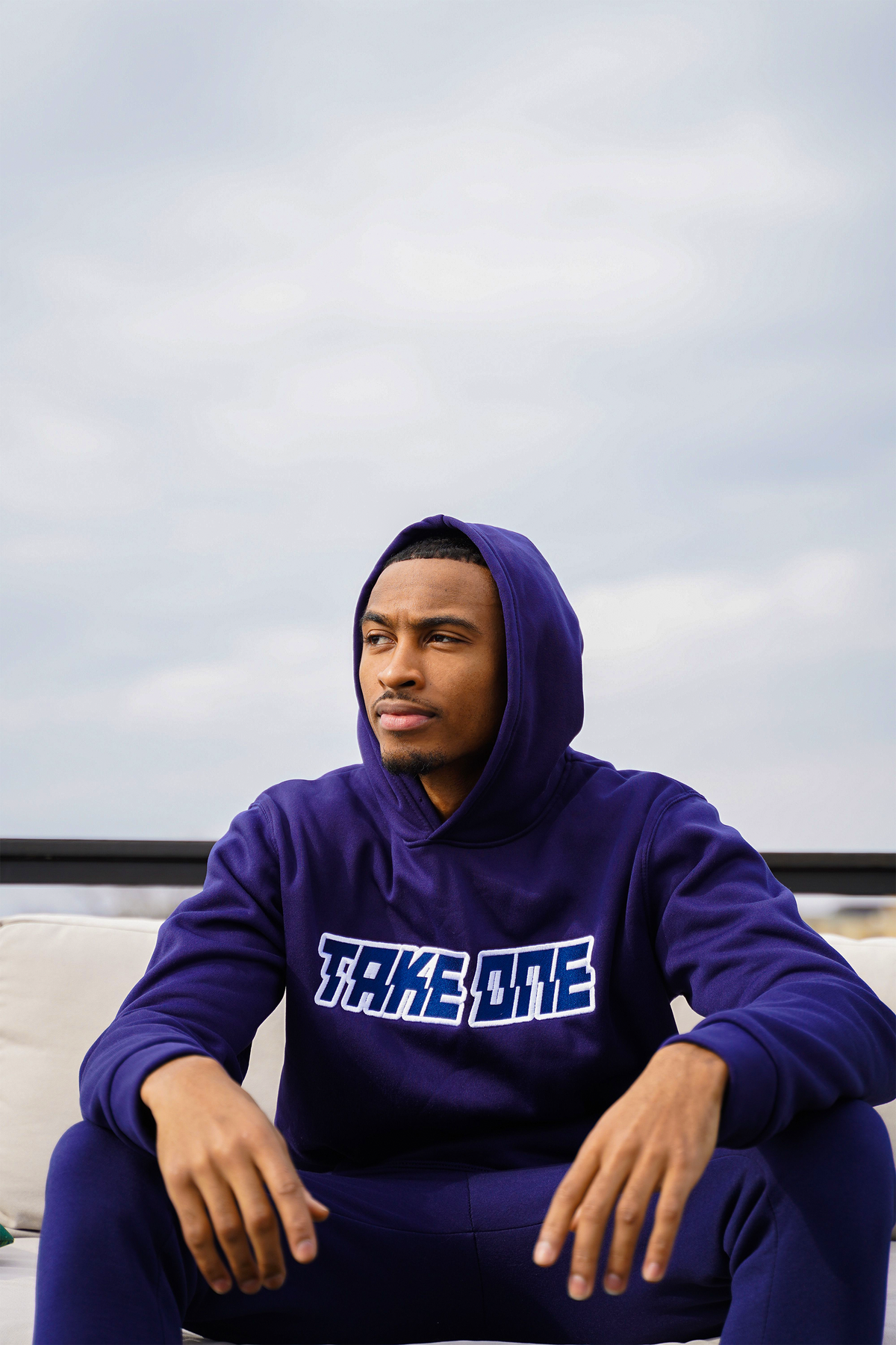 Man wearing a purple Take One Sportswear Prime Pullover Hoodie with TAKE ONE lettering while seated outdoors beneath a cloudy sky