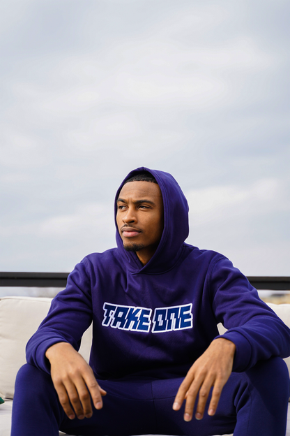 Man wearing a purple Take One Sportswear Prime Pullover Hoodie with TAKE ONE lettering while seated outdoors beneath a cloudy sky