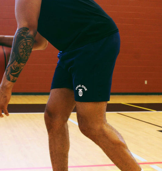 Close-up of an athlete wearing black Take One Sportswear Unlikely Winner performance basketball shorts with the Take One logo on an indoor court.