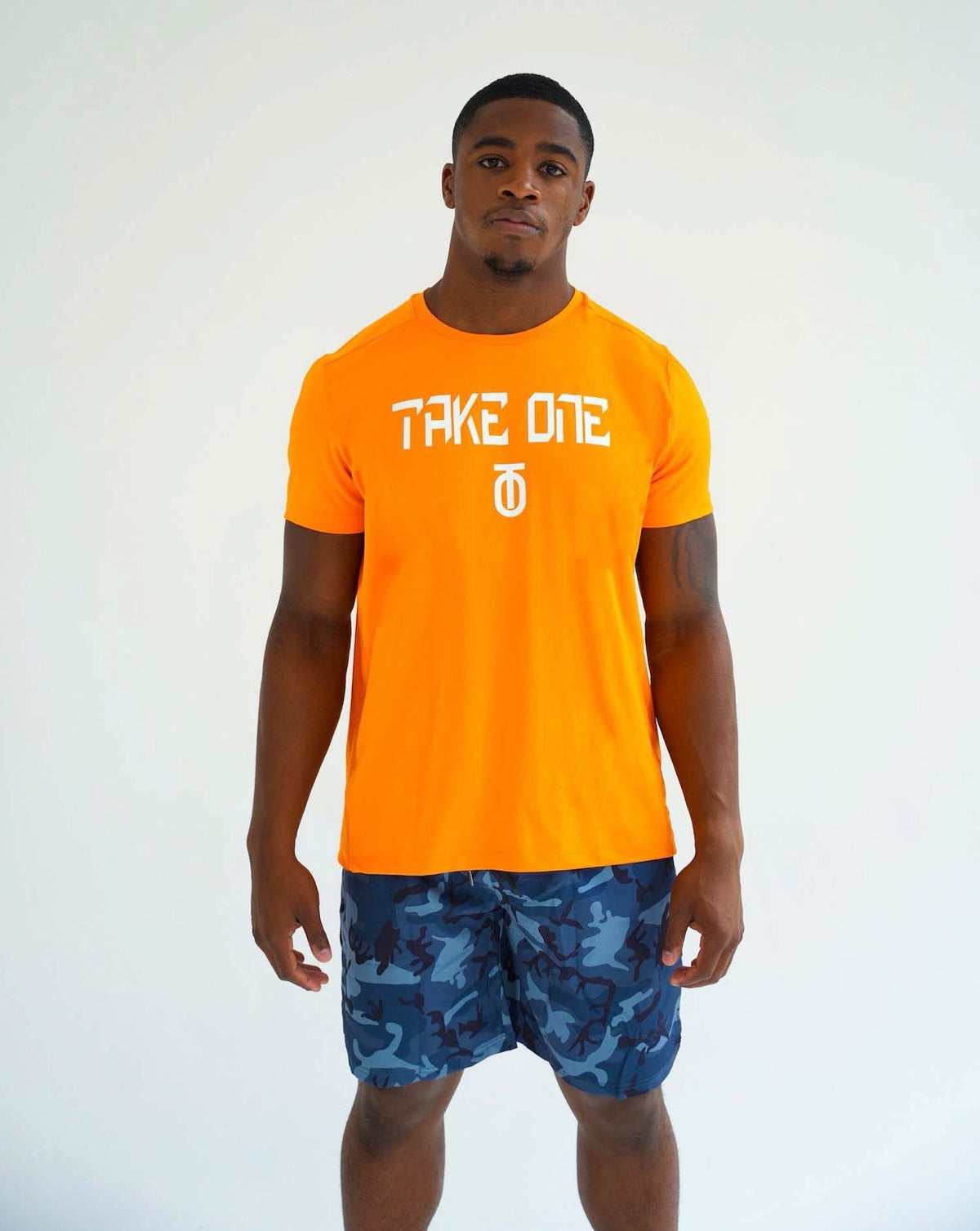 Male athlete modeling the bright orange Take One Volt FlightKnit performance T-shirt with white Take One lettering and blue patterned shorts.